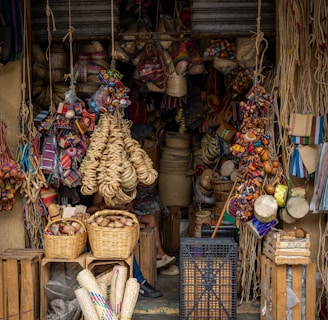 A small shop is filled with a variety of handmade goods, including woven baskets, colorful textiles, and traditional crafts. The entrance is partially obscured by hanging ropes and items such as bags and hats. Wooden crates and a plastic basket are stacked on the ground, while bright tapestries and straw items add vibrancy to the display.