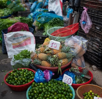A vibrant market stall displays a variety of fresh produce. There are baskets filled with limes, pineapples, cucumbers, and other leafy greens. Plastic bags, crates, and sacks are scattered around, indicating an active trading area. Price tags are visible on some products.